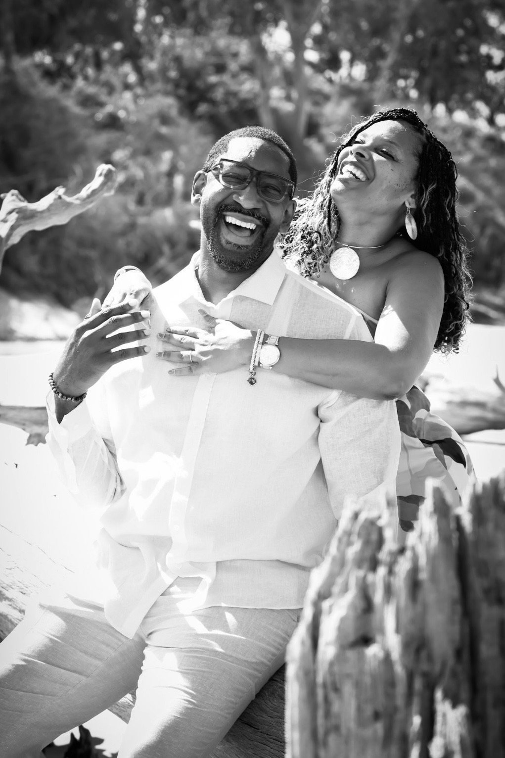 The couple laughing together on sun-bleached driftwood, her arms around his shoulders.