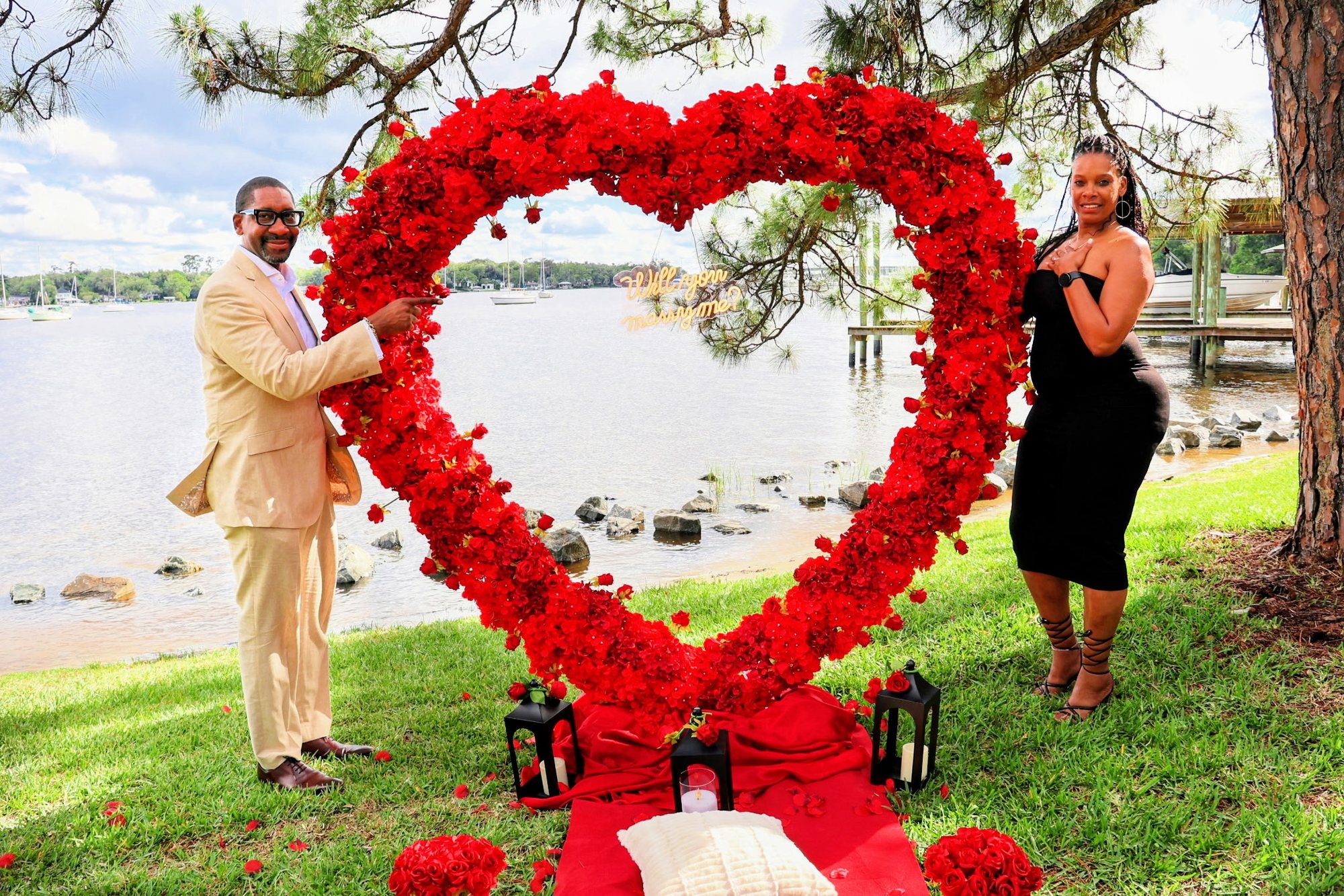 At Stinson Park, he points to a sign reading Will You Marry Me inside a heart of red roses while she covers her heart in surprise.